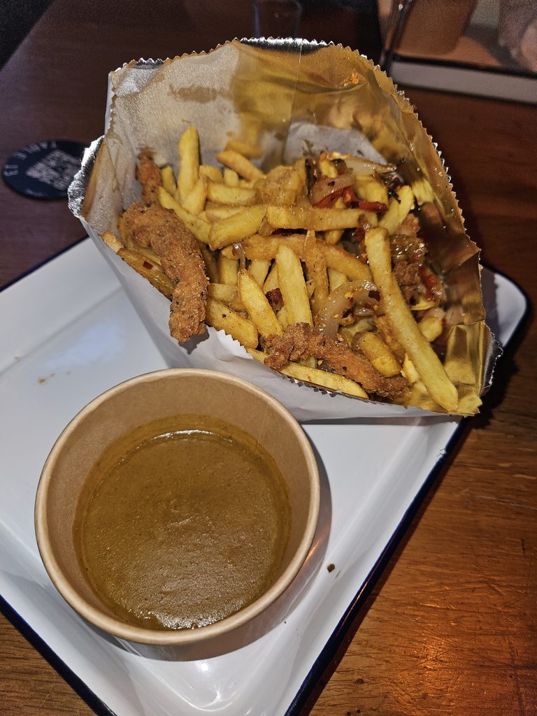 A bag of fries, fried chicken pieces, and spices with a side of curry sauce.